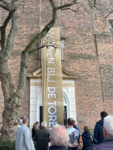 Een groep mensen staat buiten voor een historische bakstenen toren, waar een groot verticaal spandoek met de tekst 'WONEN BIJ DE TOREN' wordt onthuld. Het spandoek hangt voor een witte deur met een decoratieve omlijsting. Op de voorgrond zijn toeschouwers te zien.